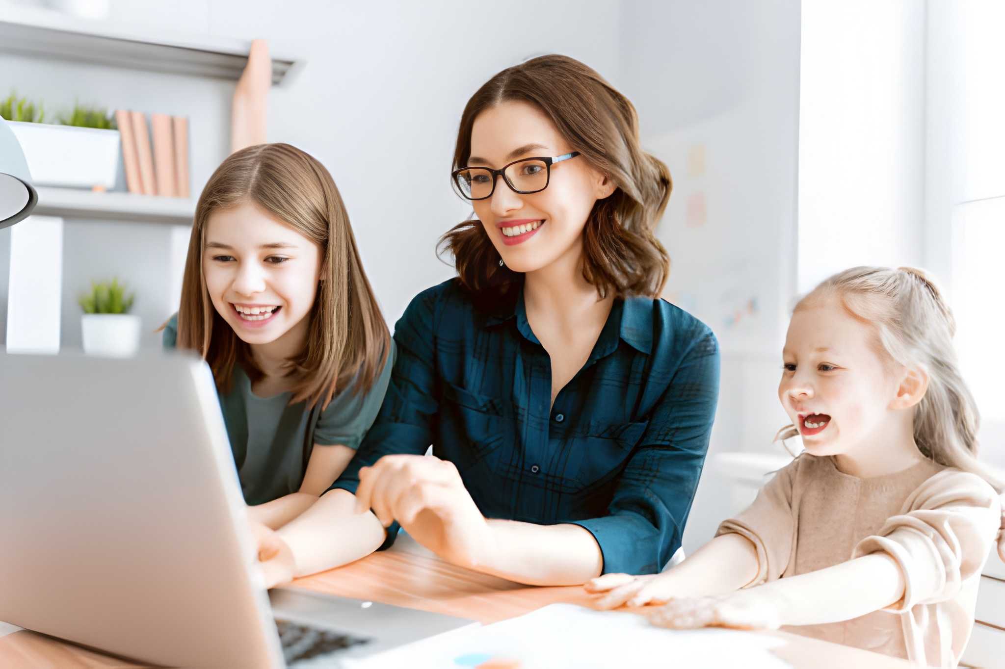 Mother helping her daughter with online schoolwork on a laptop at a home desk.